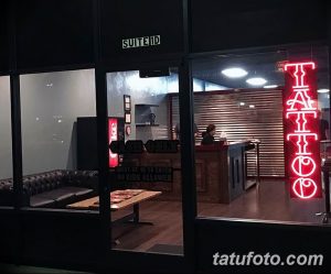Фото тату салона ночью. Яркая неоновая вывеска 'TATTOO' привлекает внимание. Внутри видно интерьер и посетителей. Современный дизайн.