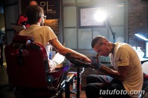 Фото процесса нанесения татуировки. Мастер в белой футболке сосредоточенно работает над татуировкой на руке клиента.