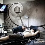 Фото процесса нанесения татуировки на мужчине в современном тату-салоне. Мастер работает над рисунком, клиент лежит на кушетке, на стене спираль.