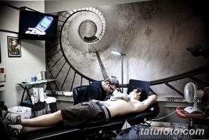 Фото процесса нанесения татуировки на мужчине в современном тату-салоне. Мастер работает над рисунком, клиент лежит на кушетке, на стене спираль.
