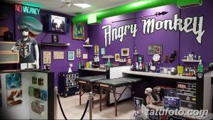 Фото интерьера тату-студии 'Angry Monkey' с фиолетовыми стенами и ярким оформлением. Видны рабочие места мастеров и элементы декора.