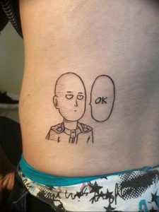 Фото тату: черно-белый рисунок Сайтамы из One-Punch Man на боку девушки. Рядом надпись "OK". Минималистичный стиль и юмористический подход.