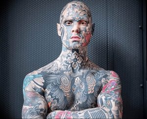 Фотография мужчины с полным боди-артом, покрывающим лицо, шею, грудь и руки. Татуировки выполнены в ярких цветах и изображают цветы, животных и другие символические мотивы.