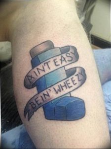 Необычная татуировка на руке с изображением ингалятора и надписи "Ain't Easy Bein' Wheezy".