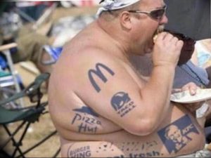 Фото тату с логотипами McDonald's, Taco Bell, Pizza Hut и KFC на груди и руках мужчины, сидящего за столом.