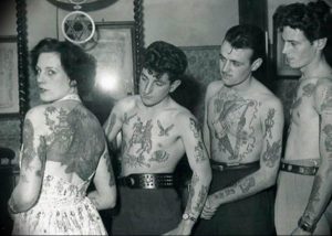 Фотография группы людей с татуировками в стиле олдскул, сделанными в начале 20-го века, демонстрирующая исторические мотивы и традиции.