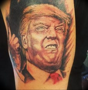 Фотография реалистичной татуировки с изображением Дональда Трампа на предплечье, выполненной в серо-коричневых тонах с высокой детализацией.