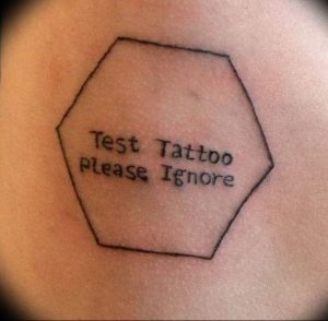Фото тату: шуточная татуировка в виде шестиугольника с надписью 'Test Tattoo Please Ignore' на руке. Черно-белый рисунок.