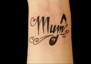Простое черно-белое фото татуировки на запястье с надписью "Mum" и изображением музыкальной ноты. Минималистичный стиль, каллиграфический шрифт.
