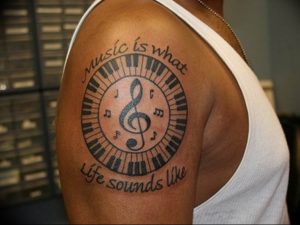 Фото татуировки на плече с изображением пианино, нот и надписи "Music is what life sounds like". Черно-белый рисунок, классический стиль.