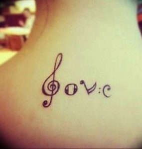 Фото тату на ключице с надписью 'Love' и скрипичным ключом. Минималистичный, женственный рисунок татуировки в черном цвете.