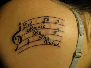 Фото тату на лопатке с изображением скрипичного ключа, нот и надписи 'Let Music Be The Voice'. Черно-белый рисунок татуировки.