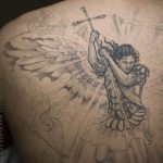 Фото тату архангела Михаила на спине, держащего крест. Детализированный рисунок в стиле реализм с использованием черных и серых оттенков.