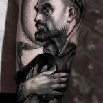 Фото тату на предплечье: реалистичный портрет монаха с нимбом и сложенными в молитве руками. Детализированный рисунок в черно-серых тонах.
