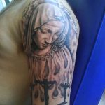 Фото тату Девы Марии с крестом на плече. Детализированный рисунок в черно-серых тонах с религиозным подтекстом и надписью.
