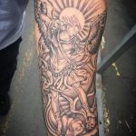 Фото тату архангела Михаила на предплечье. Детализированный рисунок в черно-серых тонах с изображением святого, сражающегося с демоном.