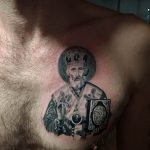 Фото тату Святого Николая Чудотворца на груди мужчины. Детализированный черно-серый рисунок с изображением святого в облачении и с книгой. Религиозная татуировка.