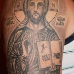 Фото тату Иисуса Христа в иконописном стиле на плече. Детализированное изображение лица с нимбом и книгой.