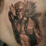Фото тату на груди с изображением Святого Николая Чудотворца в облачении епископа, выполненное в черно-серых тонах с элементами декора.