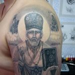 Фото тату на плече с изображением святого в облачении, держащего книгу и крест. Рисунок татуировки выполнен в реалистичном стиле с использованием темных оттенков.