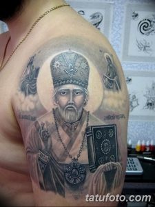 Фото тату на плече с изображением святого в облачении, держащего книгу и крест. Рисунок татуировки выполнен в реалистичном стиле с использованием темных оттенков.