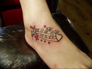 Фотография татуировки с изображением нот, сердца и надписи "Love is Music" на лодыжке. Красочный и позитивный рисунок татуировки.