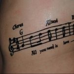 Фото тату на боку: нотная запись с надписью 'All you need is love', выполненная в черном цвете. Оригинальный рисунок татуировки.