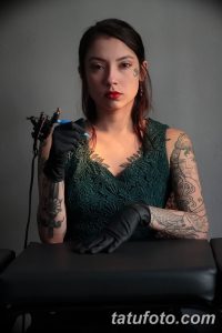 Фотография мастера тату, молодой женщины с множеством татуировок на руках и теле. Она стоит в студии, держа в руках тату-машинку.