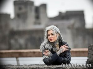 Фото девушки с серебристыми волосами и множеством татуировок на лице, шее и руках в готическом стиле.