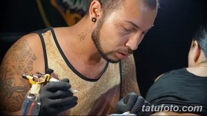 Фото процесса нанесения татуировки на руку. Мастер в майке работает над татуировкой, клиент лежит на кушетке.
