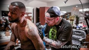 Фото процесса нанесения татуировки на плечо. Мастер в кепке и футболке работает над татуировкой, клиент сидит в кресле.