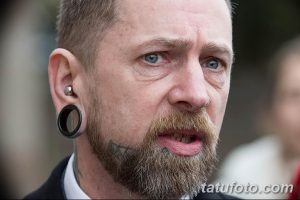 Фото мужчины с татуировкой на лице, растянутыми ушами и пирсингом. Мужчина смотрит в камеру с серьезным выражением лица.