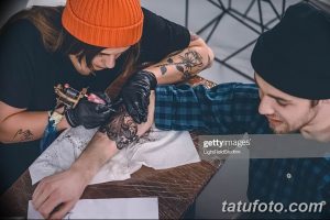 Фото тату процесса нанесения татуировки на предплечье. Мастер в черной одежде и оранжевой шапке, клиент наблюдает за работой.