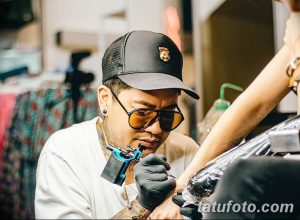 Фотография мастера тату в кепке и очках, работающего над татуировкой на руке клиента. Подчеркивает точность и мастерство.