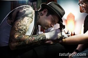 Фотография мастера тату, делающего татуировку клиенту. Видны татуировки на руках мастера и процесс нанесения рисунка.