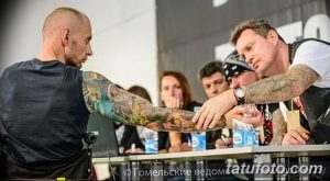 Мастер тату демонстрирует руку клиента с новой татуировкой, на фоне других людей.