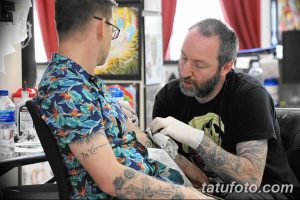 Мастер тату в черной футболке делает татуировку клиенту, сидящему в кресле.