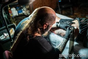 Мастер тату с длинными волосами делает татуировку на теле клиента, используя тату-машинку.