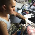 Фото процесса татуировки: мастер наносит рисунок на руку клиента. Черно-белое изображение, акцент на деталях и профессионализме.