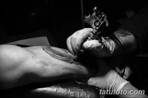Фото процесса татуировки: рука мастера с тату-машинкой наносит рисунок на плечо клиента. Черно-белое изображение, акцент на деталях и профессионализме.