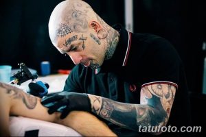 Фото тату процесса нанесения татуировки опытным мастером. Виден мастер с татуировками на теле, работающий с тату-аппаратом над рисунком на клиенте.