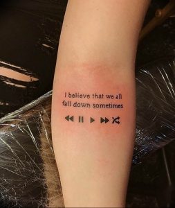 Черно-белое фото татуировки с цитатой "I believe that we all fall down sometimes" и музыкальными знаками управления плеером на предплечье.