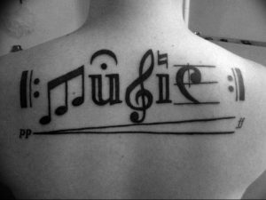 Фото тату на спине: музыкальные ноты, слово 'Music' и линии нотного стана. Оригинальный и креативный рисунок татуировки в черном цвете.
