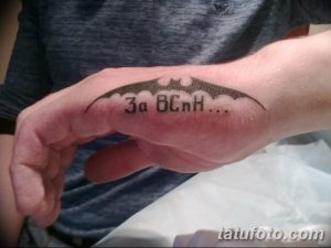 Фото тату: Изображение крыла Бэтмена и надпись 'За Всех...' на ладони. Черно-белый рисунок татуировки, выполненный в минималистичном стиле.