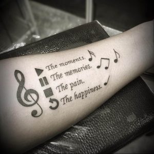 Фото тату: черно-серый рисунок ключа скрипки, нот и текста 'The moments. The memories...' на предплечье. Эмоциональный дизайн.