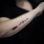 Фото тату: черная минималистичная стрела на женском предплечье. Тонкие линии, простой дизайн, популярный эскиз татуировки.