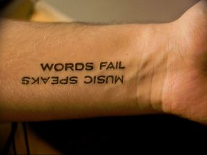 Черно-белое фото татуировки на запястье с надписью 'WORDS FAIL MUSIC SPEAKS'. Минималистичный дизайн, четкие линии, вдохновляющий смысл.