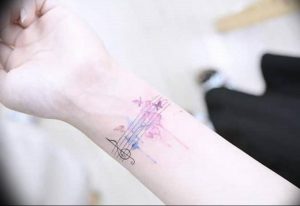 Фото тату: Нежная акварельная татуировка на руке, изображающая ноты и цветы. Пастельные тона, легкий рисунок татуировки.