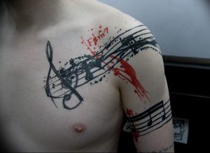 Татуировка на плече и груди, изображающая музыкальные ноты, скрипичный ключ и брызги красной краски. Оригинальный и яркий рисунок.