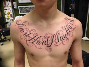 Фото тату с надписью 'Work Hard Play Hard' на груди мужчины, выполненное в стиле леттеринг с декоративными элементами.